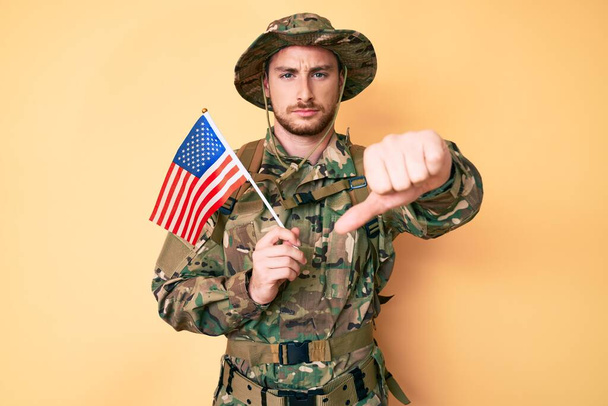 Joven hombre caucásico con camuflaje uniforme del ejército sosteniendo bandera de EE.UU. con la cara enojada, signo negativo que muestra aversión con los pulgares hacia abajo, concepto de rechazo  - Foto, Imagen