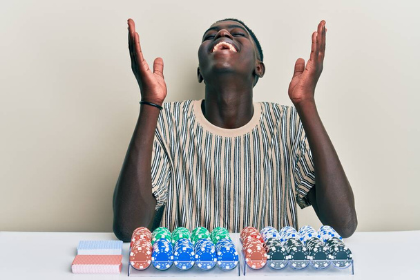 Mladý africký Američan sedí na stole s pokerovými žetony a kartami slaví šílený a šílený pro úspěch s rukama zvednutýma a zavřenýma očima křičí vzrušeně. koncept vítěze  - Fotografie, Obrázek