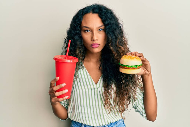 Jonge latijnse vrouw eet een smakelijke klassieke hamburger en frisdrank sceptisch en nerveus, fronsend van streek vanwege het probleem. negatieve persoon.  - Foto, afbeelding
