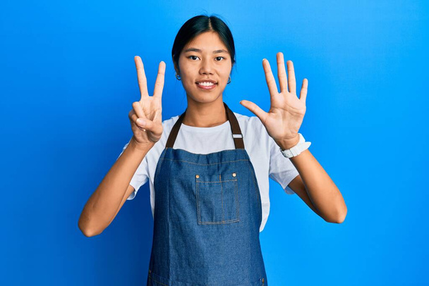Jovem chinesa vestindo avental de garçom mostrando e apontando para cima com os dedos número sete, sorrindo confiante e feliz.  - Foto, Imagem