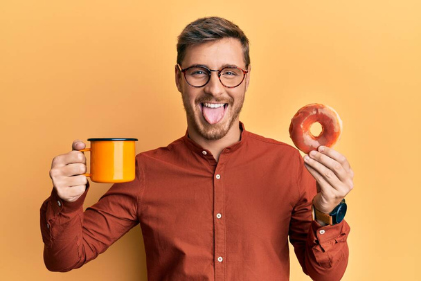 Yakışıklı beyaz adam donut yiyor ve kahve içiyor. Dilini komik bir ifadeyle dışarı çıkartıyor..  - Fotoğraf, Görsel