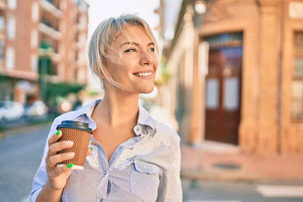 Mutlu gülümseyen ve içen genç sarışın kadın şehirde kahveyi alıp götürüyor.. - Fotoğraf, Görsel