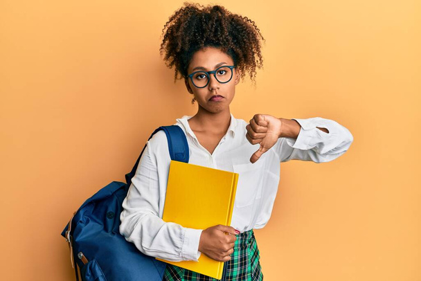 Afro saçlı güzel bir Afro-Amerikan kadını okul çantası takıyor ve elinde kızgın yüzlü kitaplar tutuyor. Negatif işaretler baş parmakları aşağıda, reddedilme kavramından hoşlanmadığını gösteriyor.  - Fotoğraf, Görsel