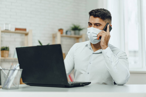 Handsome man businessman in medical mask using his laptop while talking on his cellphone - 写真・画像