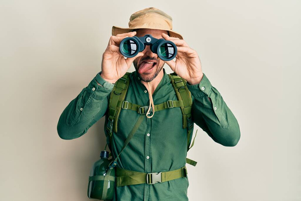 Pohledný muž s vousy nosí průzkumný klobouk dívá dalekohledem vyčnívající jazyk šťastný s legračním výrazem.  - Fotografie, Obrázek
