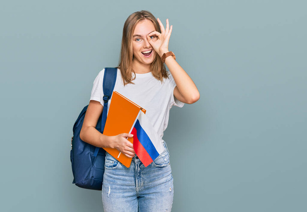 Krásná mladá blondýnka žena výměna student držení rusky vlajka usměvavý šťastný dělá ok znamení s rukou na oku dívá skrz prsty  - Fotografie, Obrázek