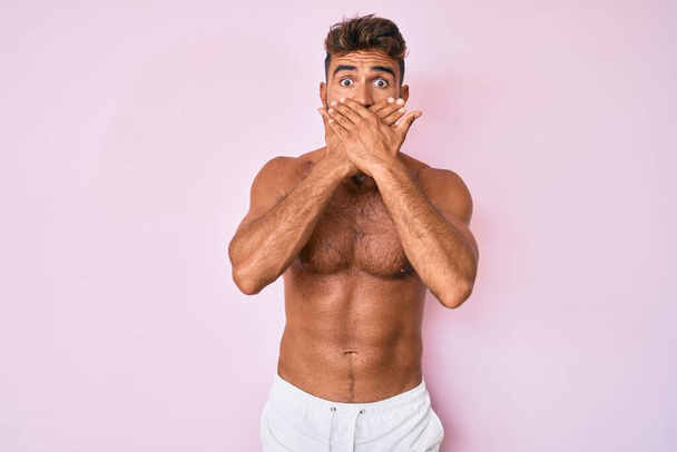 Νεαρός Ισπανός στέκεται γυμνός σοκαρισμένος καλύπτοντας το στόμα με τα χέρια του κατά λάθος. μυστική έννοια.  - Φωτογραφία, εικόνα
