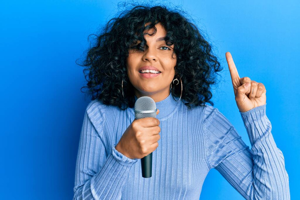 Junge hispanische Frau singt Lied mit Mikrofon lächelnd mit einer Idee oder Frage Zeigefinger mit glücklichem Gesicht, Nummer eins  - Foto, Bild