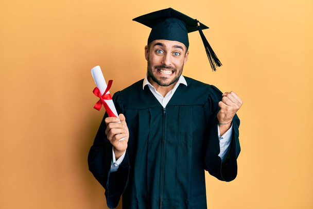 Mezuniyet cüppesi giymiş genç İspanyol adam diplomasını tutuyor gururla bağırıyor, zaferi kutluyor ve kollarını kaldırarak çok heyecanlı bir şekilde başarıyı kutluyor.  - Fotoğraf, Görsel