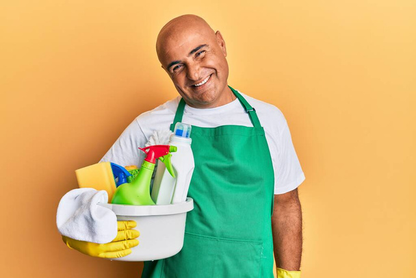 Maturo uomo del Medio Oriente indossa grembiule più pulito con prodotti per la pulizia dall'aspetto positivo e felice e sorridente con un sorriso fiducioso che mostra i denti  - Foto, immagini