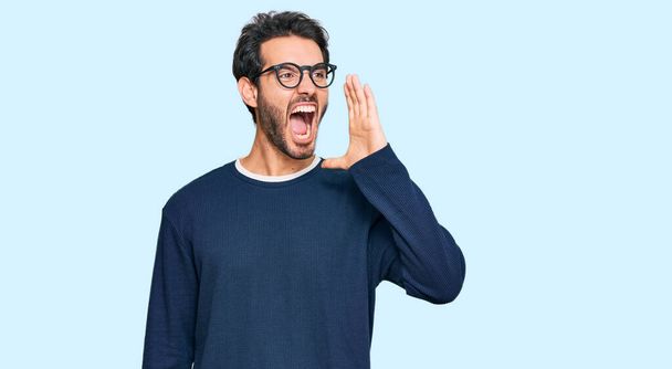 Young hispanic man wearing casual clothes and glasses shouting and screaming loud to side with hand on mouth. communication concept.  - Foto, Imagen