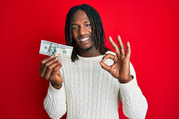 African american man with braids holding 100 dollars banknote doing ok sign with fingers, smiling friendly gesturing excellent symbol  - Foto, imagen