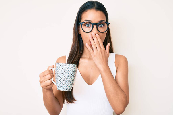 Joven hermosa chica latina con gafas sosteniendo café cubriendo la boca con la mano, conmocionada y temerosa de equivocarse. expresión sorprendida  - Foto, imagen
