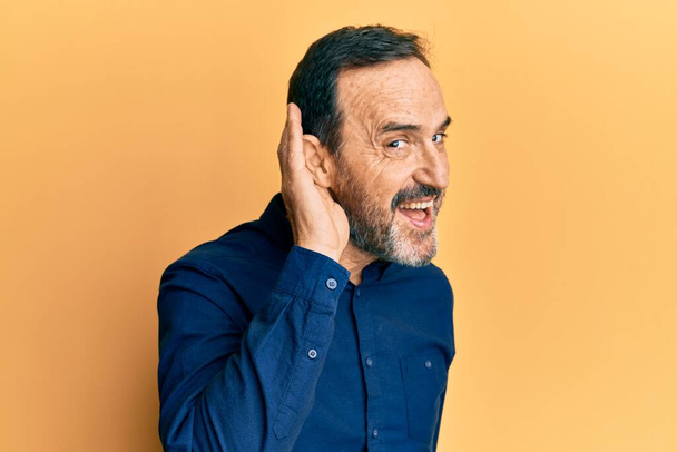 Középkorú spanyol férfi alkalmi ruhát visel, kéztől fülig mosolyog, hallgatja a pletykákat vagy pletykákat. süketség fogalma.  - Fotó, kép