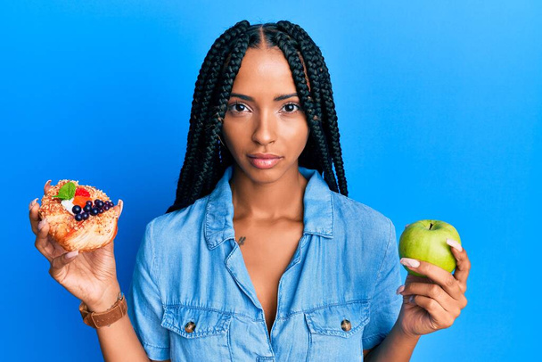 Beautiful hispanic woman holding pastry and healthy green apple relaxed with serious expression on face. simple and natural looking at the camera.  - Foto, Imagen