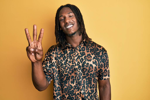 homme afro-américain avec des tresses portant léopard imprimé animal chemise montrant et pointant vers le haut avec les doigts numéro trois tout en souriant confiant et heureux.  - Photo, image