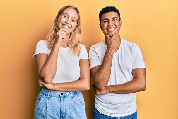 Jeune couple interracial portant un t-shirt blanc décontracté regardant avec confiance la caméra avec le sourire avec les bras croisés et la main levée sur le menton. penser positivement.  - Photo, image
