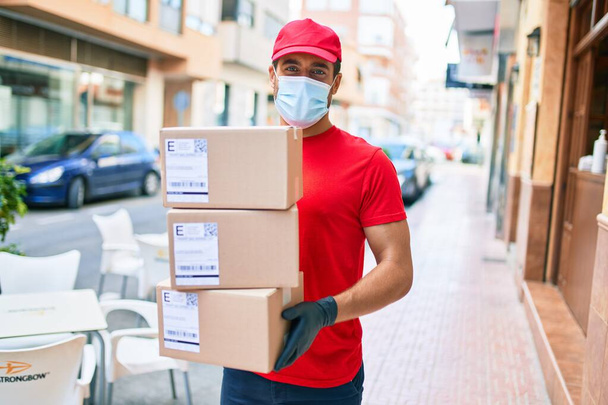 Giovane uomo di consegna indossa uniforme e protezione coronavirus maschera medica contenente pacchetti di cartone in strada città. - Foto, immagini