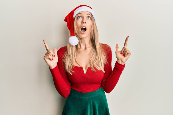 Красива біла жінка в різдвяному костюмі і капелюсі здивована і здивована, дивлячись вгору і вказуючи пальцями і піднявши руки
.  - Фото, зображення