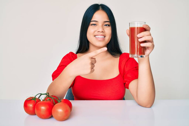 Young beautiful asian girl drinking glass of healthy tomato juice smiling happy pointing with hand and finger  - Foto, Imagem