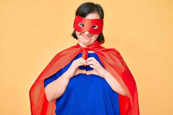 Брюнетка с синдромом Дауна в костюме супергероя улыбается в любви, показывая символ сердца и форму руками. романтическая концепция.  - Фото, изображение