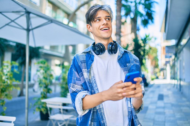 Молодой латиноамериканец улыбается счастливо, используя смартфон и наушники в городе. - Фото, изображение