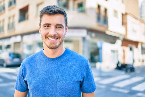 Νεαρός καυκάσιος άντρας χαμογελάει ευτυχισμένος περπατώντας στην πόλη. - Φωτογραφία, εικόνα