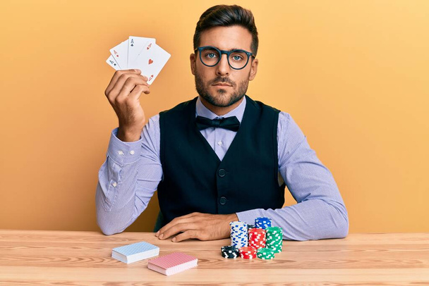Hombre guapo croupier hispano sentado en la mesa con fichas de póquer y tarjetas de pensamiento actitud y expresión sobria buscando confianza en sí mismo  - Foto, Imagen