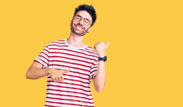 Fiatal spanyol férfi alkalmi ruhát visel, hátrafelé mutatva kezével és hüvelykujjával, mosolyogva magabiztosan.  - Fotó, kép