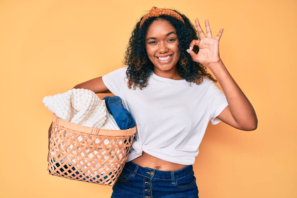 Joven mujer afroamericana sosteniendo cesta de la ropa haciendo signo bien con los dedos, sonriendo gesto amistoso excelente símbolo  - Foto, Imagen