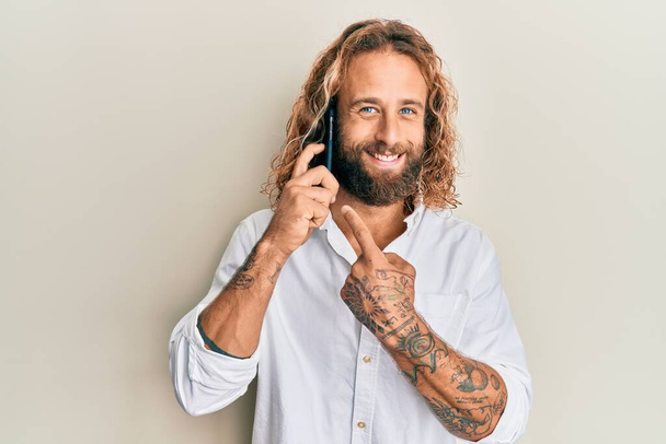 Handsome man with beard and long hair having conversation talking on the smartphone smiling happy pointing with hand and finger  - Fotografie, Obrázek