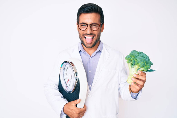 Young hispanic man as nutritionist doctor holding weighing machine and broccoli sticking tongue out happy with funny expression.  - Φωτογραφία, εικόνα