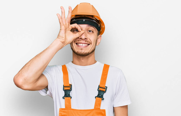 Joven hispano vistiendo uniforme de manitas y sombrero de seguridad sonriendo feliz haciendo señal de bien con la mano en el ojo mirando a través de los dedos  - Foto, imagen