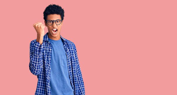 Young african american man wearing casual clothes and glasses angry and mad raising fist frustrated and furious while shouting with anger. rage and aggressive concept.  - Foto, imagen