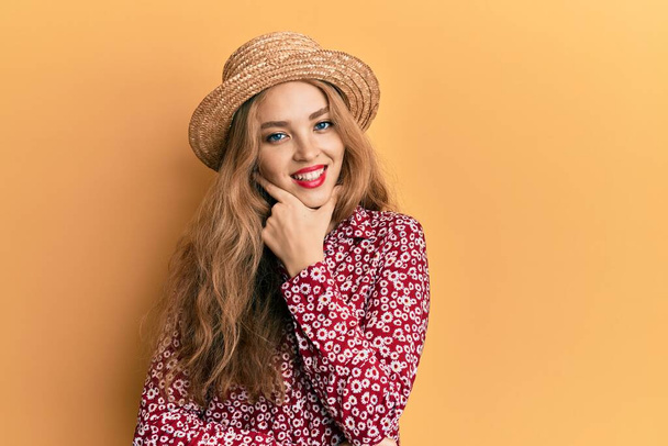 Beautiful blonde caucasian woman wearing summer hat looking confident at the camera smiling with crossed arms and hand raised on chin. thinking positive.  - Photo, Image