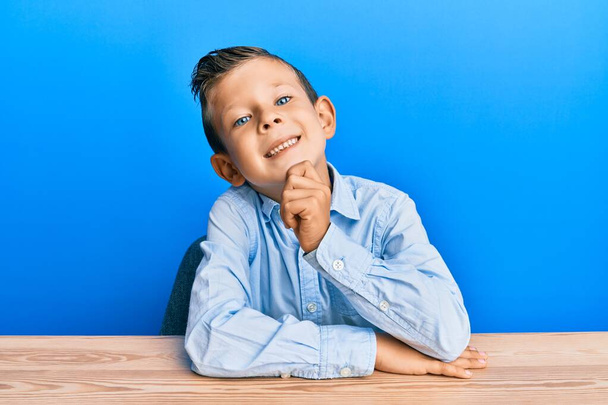 Schattige blanke jongen draagt casual kleding zittend op de tafel glimlachend zelfverzekerd kijkend naar de camera met gekruiste armen en hand op kin. positief denken.  - Foto, afbeelding