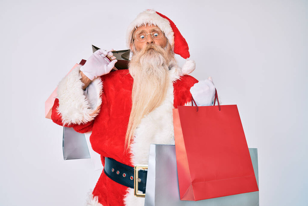 Velho homem sênior com cabelos grisalhos e barba comprida vestindo traje de Papai Noel segurando sacos de compras fazendo cara de peixe com boca e olhos cintilantes, louco e cômico.  - Foto, Imagem