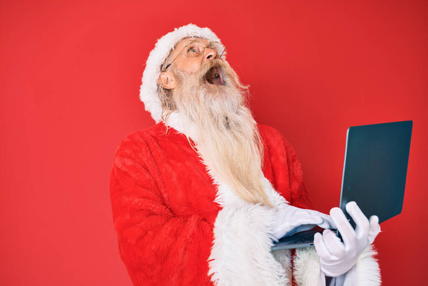 Velho homem sênior com cabelos grisalhos e barba longa vestindo traje de Papai Noel usando laptop irritado e louco gritando frustrado e furioso, gritando com raiva olhando para cima.  - Foto, Imagem