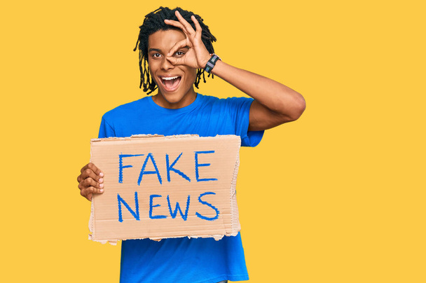 Jovem afro-americano segurando falso banner de notícias sorrindo feliz fazendo ok sinal com a mão no olho olhando através dos dedos
  - Foto, Imagem