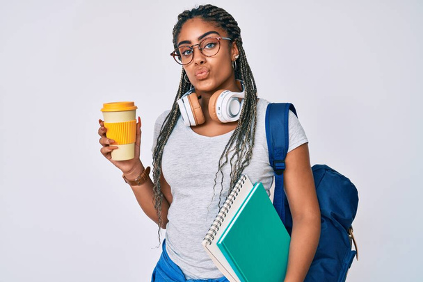 Fiatal afro-amerikai nő copfokkal, aki diákhátizsákot visel, kávét iszik, miközben a kamerába néz, és egy csókot fúj, hogy kedves és szexi legyen. szerelem kifejezés.  - Fotó, kép