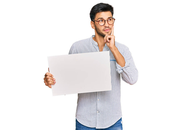 Young handsome man holding blank empty banner serious face thinking about question with hand on chin, thoughtful about confusing idea  - Photo, Image