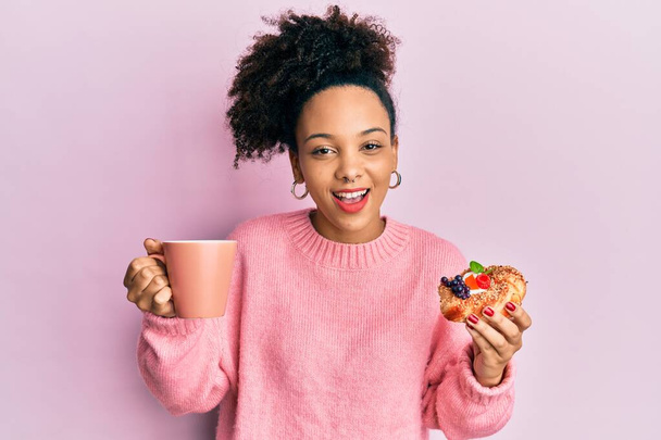 Chica afroamericana joven bebiendo una taza de café y comiendo dulce sonrisa y riendo en voz alta porque broma loca divertida.  - Foto, Imagen