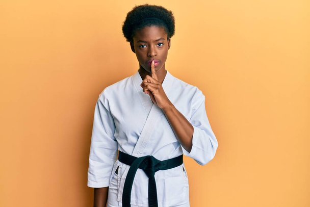 Jovem afro-americana vestindo kimono de karatê e faixa preta pedindo para ficar quieta com o dedo nos lábios. silêncio e conceito secreto.  - Foto, Imagem