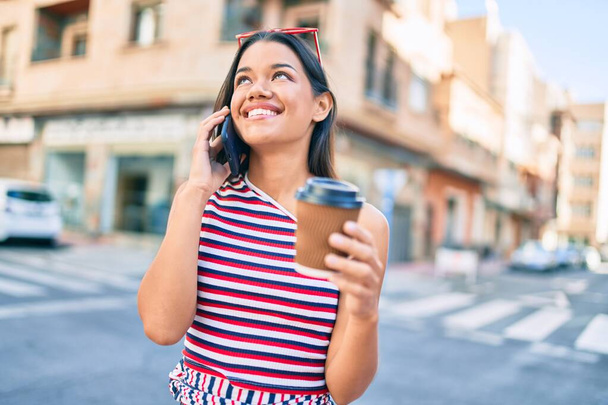Jeune fille latine souriant heureux parler sur le smartphone et boire du café à emporter à la ville. - Photo, image