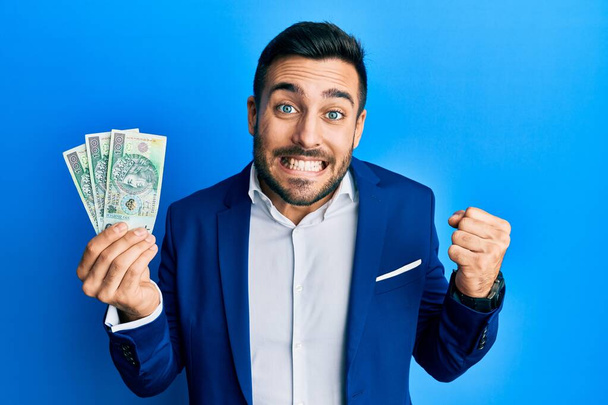 Junger hispanischer Geschäftsmann im Businessanzug hält polnische Zloty-Banknoten in der Hand, schreit stolz und feiert Sieg und Erfolg sehr aufgeregt mit erhobenem Arm  - Foto, Bild