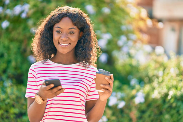 jong Afrikaans amerikaans meisje met behulp van smartphone drinken koffie in het park. - Foto, afbeelding