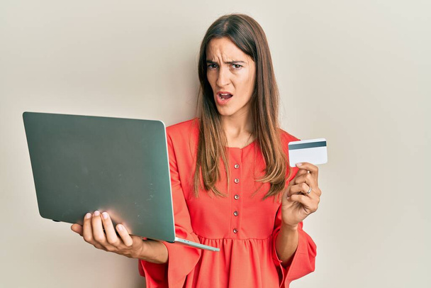 Young beautiful woman holding laptop and credit card in shock face, looking skeptical and sarcastic, surprised with open mouth  - Фото, зображення