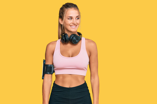 Young blonde woman wearing gym clothes and using headphones looking away to side with smile on face, natural expression. laughing confident.  - Photo, Image