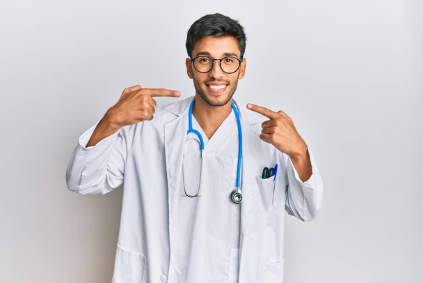 Молодой красивый мужчина в докторской форме и стетоскопе весело улыбается, показывая и показывая пальцами зубы и рот. концепция стоматологического здоровья.  - Фото, изображение