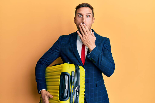 Hombre guapo con barba llevando traje de negocios sosteniendo la maleta en viaje de trabajo cubriendo la boca con la mano, sorprendido y temeroso de equivocarse. expresión sorprendida  - Foto, imagen